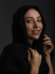 Young muslim female model in casual clothes and traditional black hijab smiling and looking at camera over dark gray background, copy space