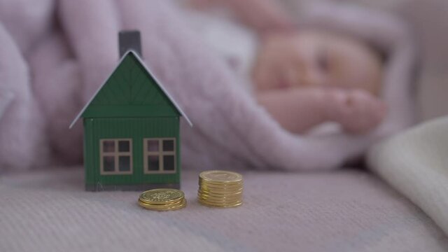 Coins and toy house on bed with blurred infant sleeping at background lying in cozy bed. Concept of newborn care and home purchase