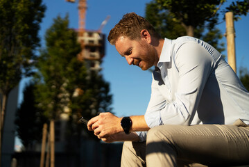 Young stylish businessman sitting on the beanch outdoors. Portrait of handsome man using the phone.