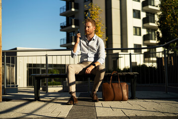 Young stylish businessman sitting on the beanch outdoors. Portrait of handsome man using the phone.