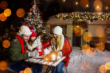 Family, winter holidays and celebration concept - happy mother, father and little daughter with drinks at Christmas eve, New year sitting at the table with decorated trailer on background