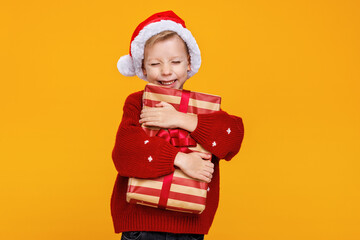 Cute happy boy in santa hat hugging big Christmas gift box tightly and closing his eyes with joy