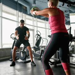 Fototapeta na wymiar Middle-aged woman repeating after personal trainer squats with gym ball
