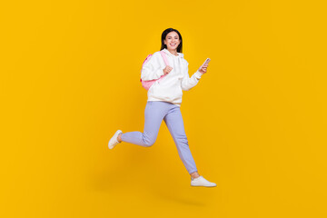 Fototapeta na wymiar Full body photo of youth lady jump use app smart phone hold rucksack isolated over shine yellow color background