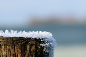 Top of a wooden fence pole covered with ice crystals, lit by morning sun. Concepts of frosty winter season, cold temperature, hoarfrost. Macro shot with background blur and copy space.
