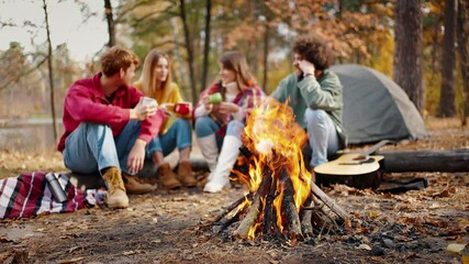 Friends smiling, enjoying tea on picnic, sitting on log in autumn wood near river. Tent, burning campfire. Slow motion, blurred background