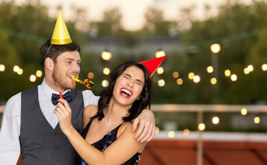birthday, celebration and holidays concept - happy couple with party blowers and caps having fun at rooftop party over lights on background