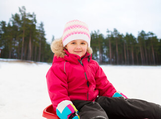 childhood, sledging and season concept - happy little girl sitting in sled outdoors in winter over pine forest background