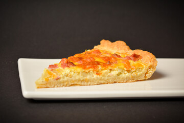 Side view of piece of traditional French pie quiche Lorraine with ham, onion and eggs. Backed custard dough served on white plate. Classic recipe. Black background.
