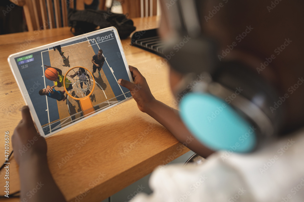 Wall mural African american boy wearing headphones at home watching basketball game on tablet