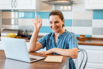 Happy smiling young beautiful woman talking on video call, waving hand greetings using laptop at home in kitchen interior. Woman freelancer working remotely or study at home e-learning. 