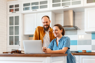 Smiling young married couple talking via video call using laptop at home in kitchen interior. Woman and man happy to hear good news from e-mail in laptop. Video calls communication. 