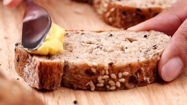 Women holding sliced multigrain bread on wooden table and using butter knife spreading tasty butter cream on fresh bread with seed.