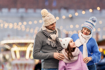 family, people and winter clothes concept - happy mother, father and little daughter in knitted hats and scarves outdoors over christmas market and lights background