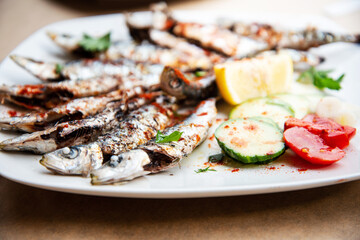 Fresh Grilled sardines served with fresh tomatoes, cucumbers slices and lemon on white plate for mediterranean healthy meal