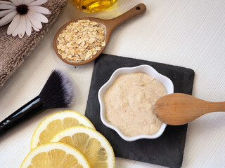 DIY face mask in white ceramic bowl, brush, wooden spoon, ceramic spoon with oat flakes, lemon slices, flower on a white background. Flat lay