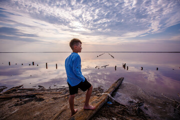 Rear view of a cute teenage boy in summer clothes standing on an amazing pink lake with salt and healing mud. Sunset sky.