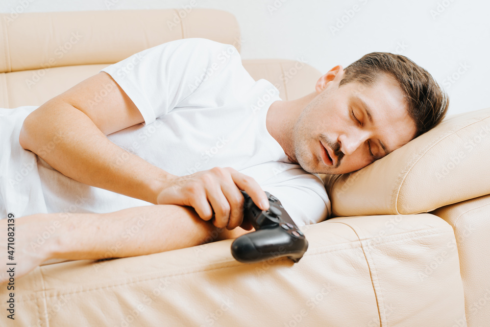 Wall mural tired young man holding wireless gamepad controller and sleeping on sofa at home. adult guy gamer pl