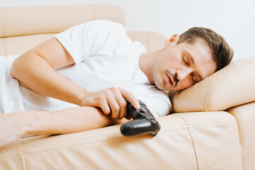 Tired young man holding wireless gamepad controller and sleeping on sofa at home. Adult guy gamer...