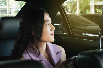 Young Asian beautiful woman sitting of back seat inside of the car and looking outside through car window while traffic. Beautiful female with a little smile chatting with friend on her mobile phone.