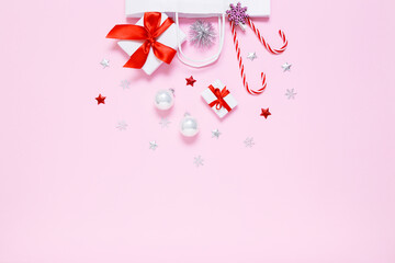 Christmas, New Year composition. White paper bag with presents, sweet candy canes, holiday decorations, silver balls, glitter confetti stars on pastel pink background. Flat lay, copy space.