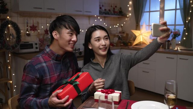 Happy asian korean couple exchanging gifts with each other at home in decorated kitchen with warm light. cute lovers taking self photo with present box during xmas eve celebration