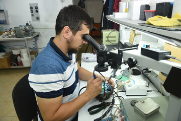 A male repairman in the workshop looks at the microcircuit of an electrical device through a microscope while doing electrical soldering of equipment