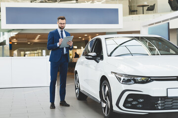 Young handsome caucasian shop assistant in formal clothes holding clipboard checking car options...