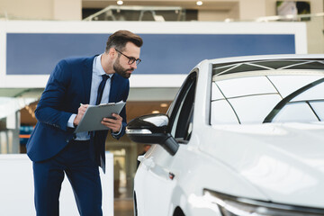 Successful handsome caucasian shop assistant in formal clothes holding clipboard writing checking car options information at automobile dealer shop store.