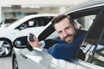 Happy car owner. Successful smiling caucasian businessman holding car keys after buying automobile while sitting at the wheel of a new expensive auto at dealer shop