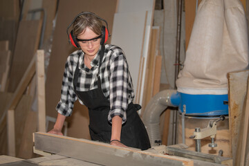 Young woman as a carpenter with safety glasses and hearing protection works on planing machine - obrazy, fototapety, plakaty