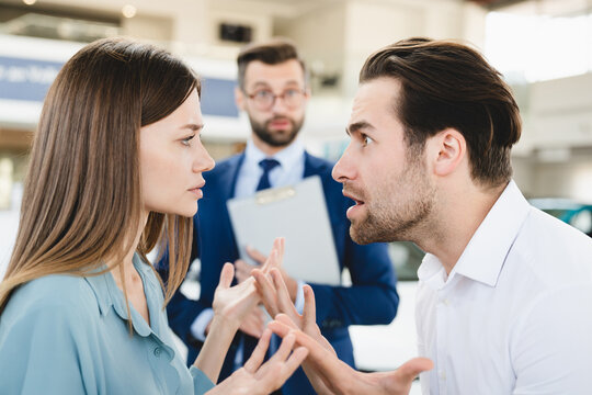Focused photo of confused male shop assistant looking at angry arguing young family caucasian couple while they are deciding which car auto to choose buy. Expensive high price.