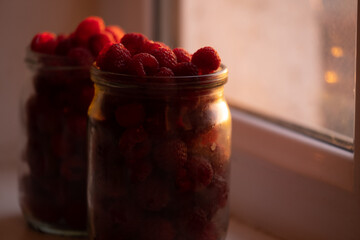 Fresh raspberries in a jar on the windowsill close-up. The rays of the sunset illuminate everything through the window with warm light.