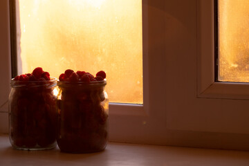 Fresh raspberries in a jar on the windowsill close-up. The rays of the sunset illuminate everything through the window with warm light.