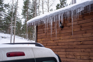 Icicles hang from the roof of a log house near which a car is carelessly parked, which can be...