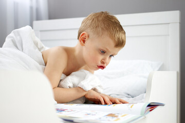 A little boy voices a book that he is looking at while lying in a crib under a blanket