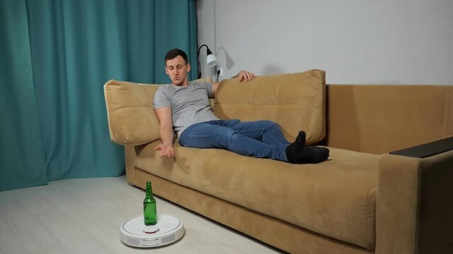 White robot vacuum cleaner delivers alcohol beer bottle to brunet short-haired man sitting on sofa in grey t-shirt, jeans in living room.