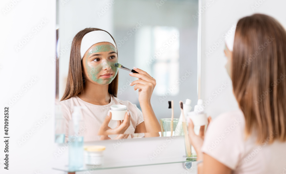 Sticker beauty, skin care and people concept - teenage girl with brush applying clay mask to face and looking in mirror at bathroom