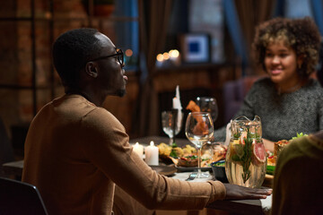 Rear view of African young man in eyeglasses sitting at dining table with his friends and praying before eating