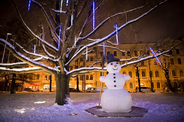 Panorama of a winter evening in the city, illuminated by evening street lights, before Christmas, New Year. Snowy streets. Trees decorated with lights. Riga, Latvia. The snowman is cute.