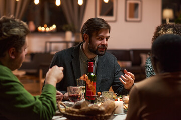 Young man sitting at dining table and talking to the people during dinner party at home