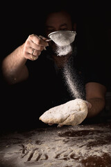 Cooking, kneading dough. Male hands sift flour onto the dough on a dark background.