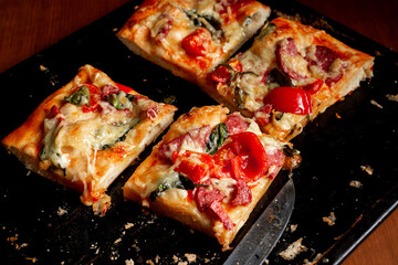 Sliced square pieces of pizza, pie, with tomatoes, sausage, cheese cooked in the oven on a black baking sheet.