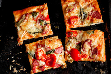 Sliced square pieces of pizza, pie, with tomatoes, sausage, cheese cooked in the oven on a black baking sheet.