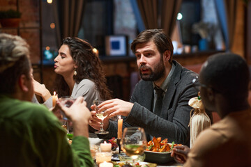 Young bearded man in suit talking to people while sitting at dining table during dinner
