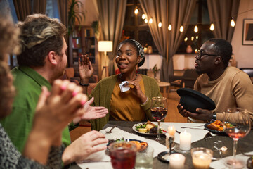 African young woman gesturing while sitting at dining table and playing the game with her friends at home