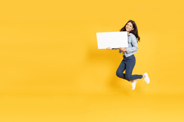 Young Asian woman showing and holding blank white billboard and jumping isolated on yellow...
