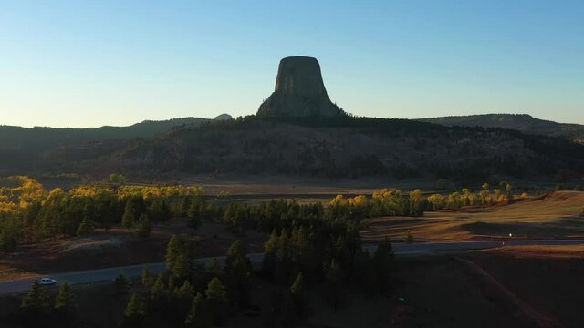 Devils Tower Butte and Cars on Road at Sunset in Autumn. Yellow Trees. Crook County. Wyoming, USA. Aerial View. Drone Flies Sideways