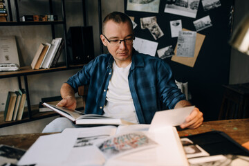 A young man with glasses at work at the table holding a notebook.