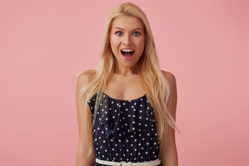 Studio Portrait of blonde female wears polka dot dress keeps mouth widely opened with surprised facial expression. isolated over pink background